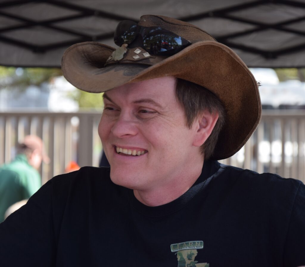 Micha Petty outdoors at a public event, wearing a wide-brimmed hat and smiling off-camera.