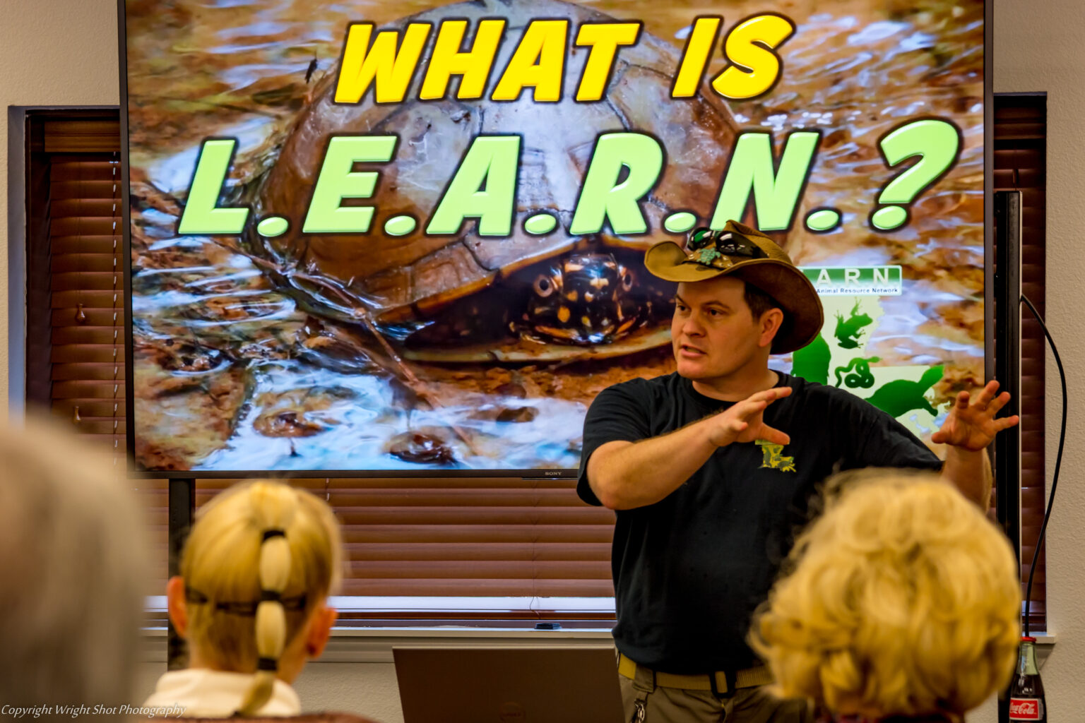 Micha Petty presenting to an audience in front of a large screen showing a turtle and the title “What is LEARN?”