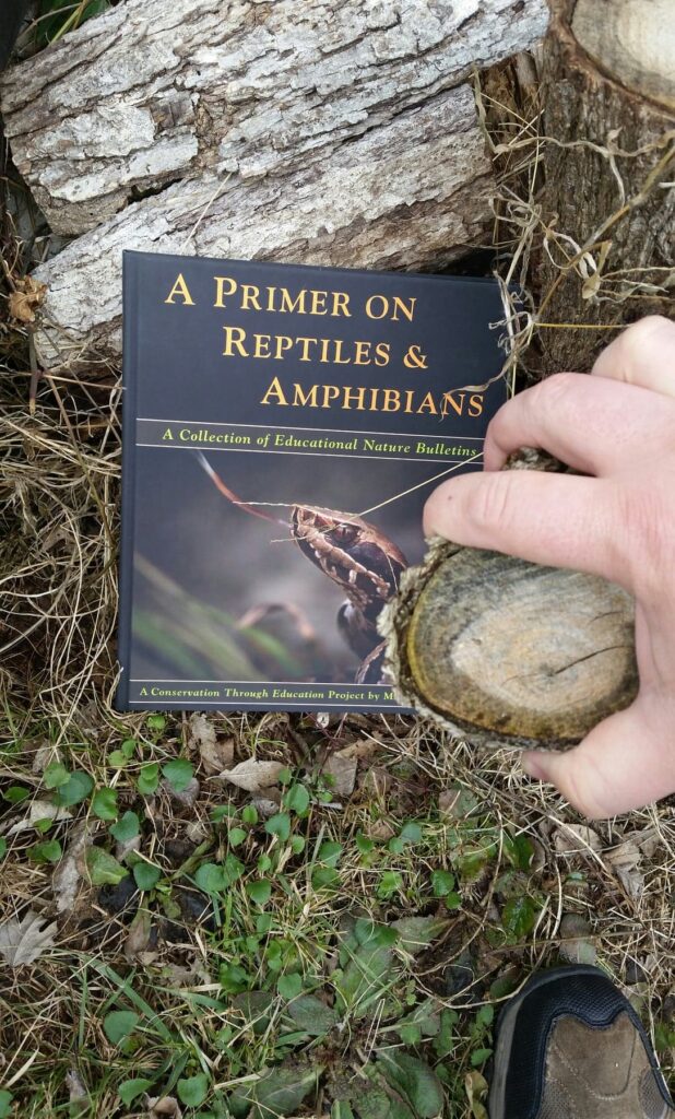 A reader holding a copy of the Primer outdoors beside a lichen-covered rock.