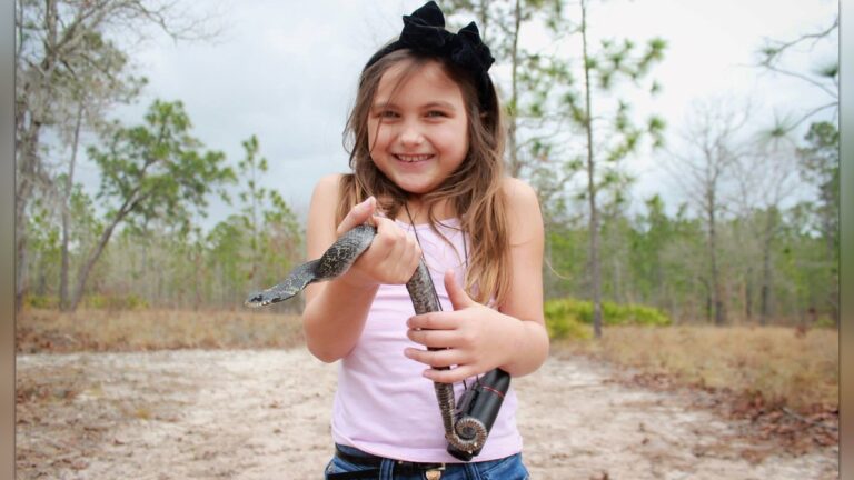 A smiling child holding a small snake outdoors in a pine woodland.