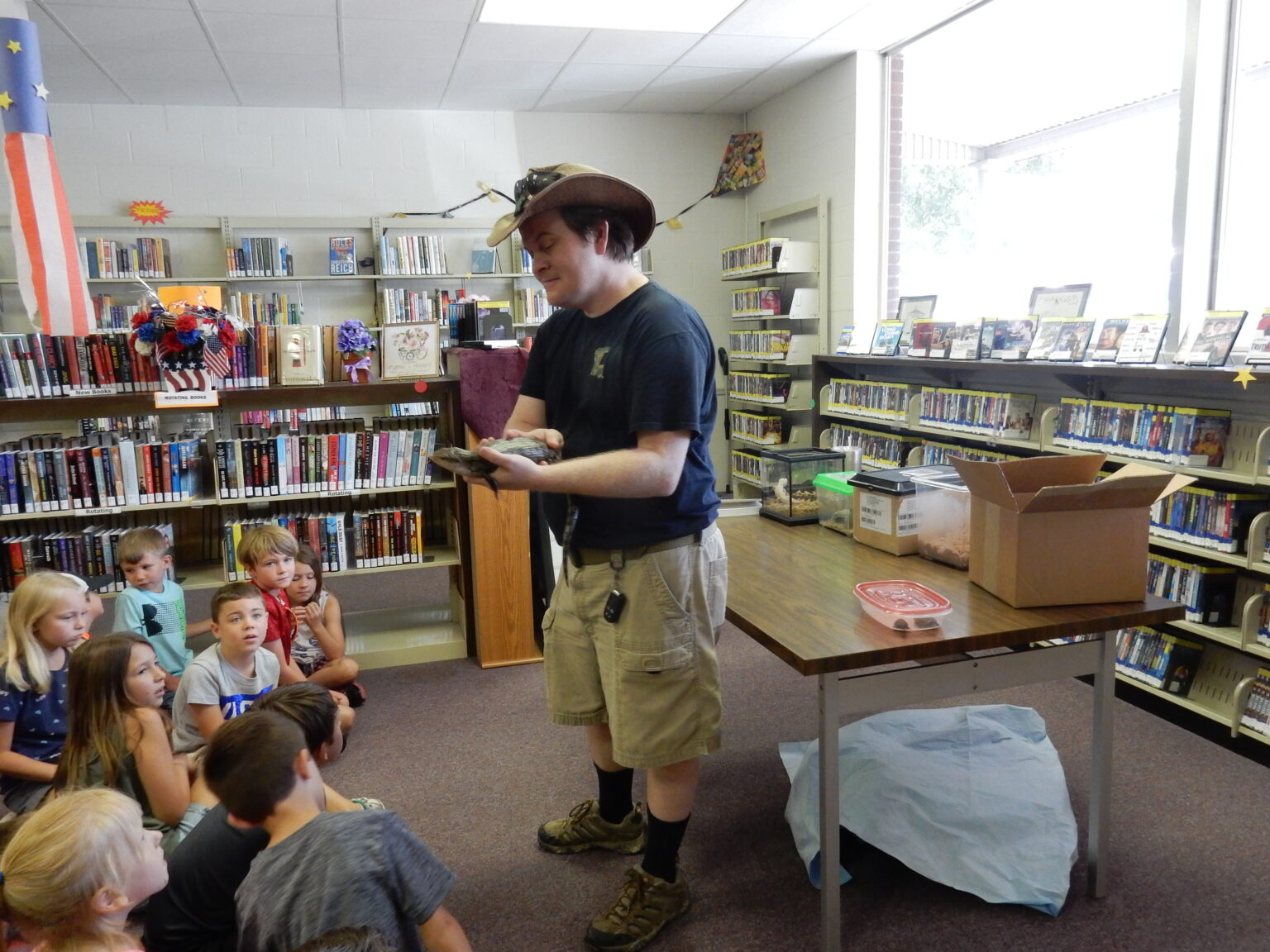 Micha Petty showing an animal to a group of children seated on the floor of a public library.